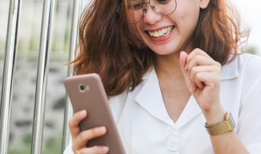 Une femme en chemise blanche rit en regardant l'écran de son smartphone