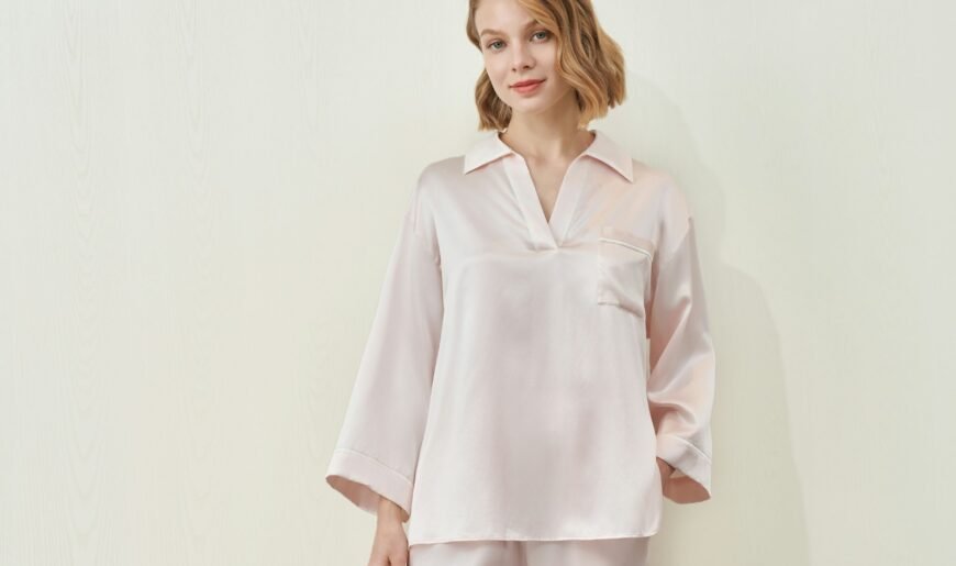 Une femme porte un ensemble pyjama en soie rose pâle, chemise et pantalon, devant un mur clair